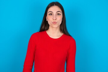 Fototapeta premium Shot of pleasant looking young brunette girl wearing red T-shirt against blue wall , pouts lips, looks at camera, Human facial expressions