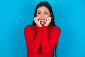 Fearful young brunette girl wearing red T-shirt against blue wall keeps hands near mouth, feels frightened and scared,  has a phobia,  Shock and frighted concept.