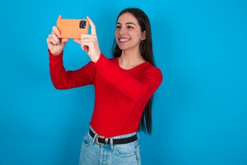 young brunette girl wearing red T-shirt against blue wall taking a selfie to post it on social...