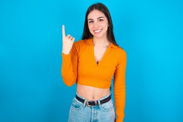 Fototapeta na wymiar young caucasian brunette girl wearing orange crop top against blue wall smiling and looking friendly, showing number one or first with hand forward, counting down