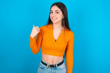 Fototapeta na wymiar young caucasian brunette girl wearing orange crop top against blue wall points away and gives advice demonstrates advertisement
