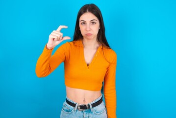 young caucasian brunette girl wearing orange crop top against blue wall purses lip and gestures with hand, shows something very little.