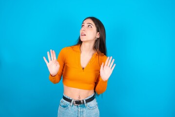 young caucasian brunette girl wearing orange crop top against blue wall keeps palms forward and looks with fright above on ceiling tries to defense herself from invisible danger opens mouth.