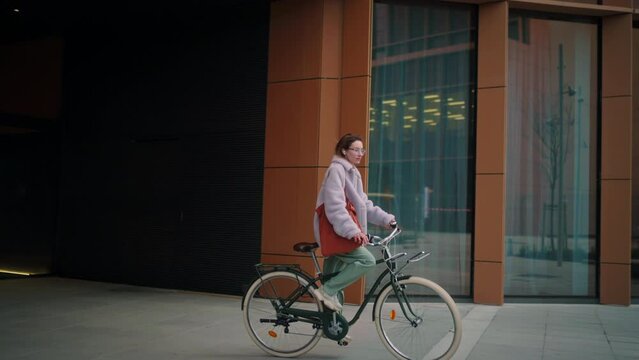 young trendy looking woman wearing eyewear cycling around the city near a modern urban business building. good vibe concept female hipster riding a green rental vintage bike