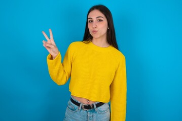 young brunette girl wearing yellow knitted sweater against blue wall makes peace gesture keeps lips folded shows v sign. Body language concept