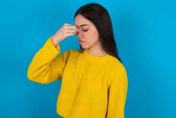 Sad young brunette girl wearing yellow knitted sweater against blue wall suffering from headache holding hand on her face