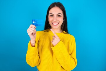 Photo portrait of young brunette girl wearing yellow knitted sweater against blue wall doing...