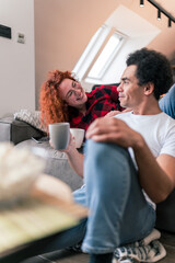Beautiful red-haired woman with hipster glasses is lying on the couch, drinking coffee and telling a funny story to her partner sitting on the floor