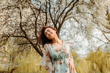 Portrait of charming pretty woman dressed flowery dress posing near apple cherry tree blossoms blooming flowers in the garden park in early spring nature. Fashion, girl model with black hair
