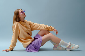 Fashionable confident girl wearing purple sunglasses, pleated mini skirt, yellow hoodie, socks, high top colorful sneakers, sitting, posing on blue background. Full-length studio portrait. Copy space - obrazy, fototapety, plakaty