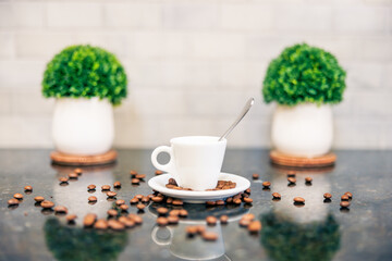 Espresso cup and saucer with spoon and dark roasted coffee beans with modern counter and white subway tile reflection in cafe bar kitchen with green plants
