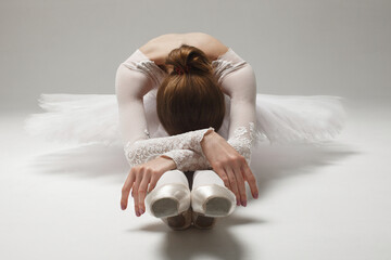 beautiful ballerina in white ballet clothing sitting on floor bent over, studio shot - obrazy, fototapety, plakaty