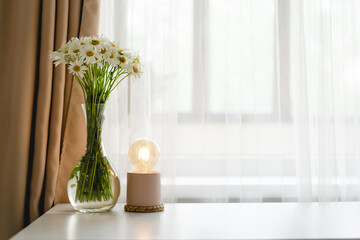 Workplace with flowers and decorative small lamp on the table against window. Workplace at home