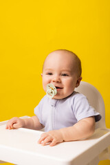 cute baby with a pacifier in his mouth in a high chair on a yellow background. Baby food, space for text