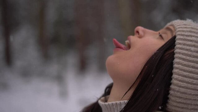 Cute teenage girl catching snowflakes with her tongue outdoors, beige hat, Austria forest, smiling