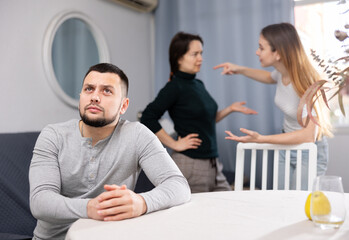 Two women quarrel while sad man sits at table at home