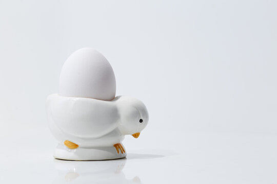 Selective focus view of boiled egg in cute ceramic chick holder on plain white shiny background
