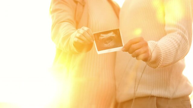 Unrecognised of a pregnant woman with her husband are holding an ultrasound scan photo of the unborn child outdoors on nature in sunlights. Happy motherhood and parenthood concept.
