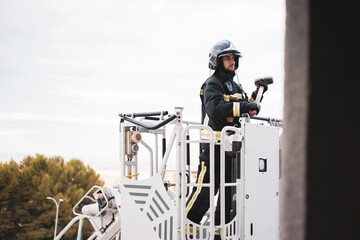 A young and uniformed firefighter is mounted in the basket of a crane truck, the man carries in his hands a sledgehammer to break a window of the building.