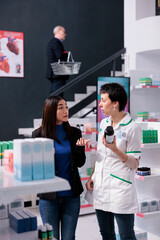 Young asian woman buying vitamin and consulting with pharmacy worker. Medical retail store employee holding multivitamin pills bottle and answering customer questions about food additive
