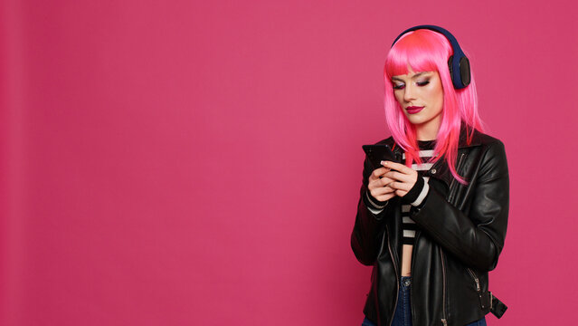 Cheerful confident adult with pink hair texting messages, listening to music on headset. Cool stylish woman having fun with mp3 songs, using social media online app on smartphone.
