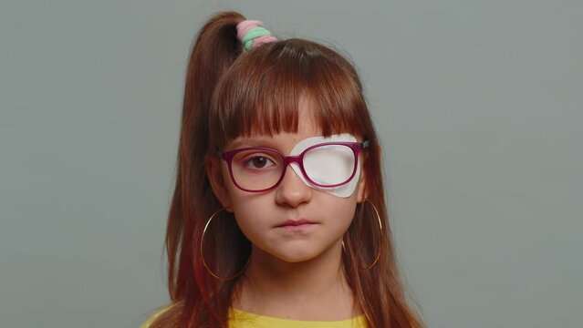 Sick ill preteen girl looking at camera having protective bandage plaster patch scratch on one eye. Injury, trauma, blind. Child kid wearing glasses at gray background. Medical eyesight vision therapy