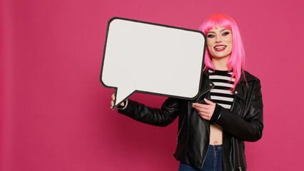 Beautiful trendy person holding isolated speech bubble in studio, using blank copyspace cardboard to create advertisement. Young cheerful model showing carton ad on camera, pink hairstyle.