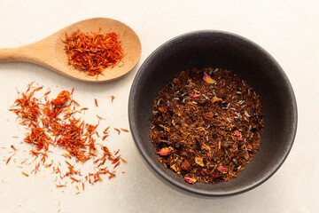 Selective focus high angle view of orange and brown coloured dried spices and leaves in black bowl and wooden spoon set on plain pale background