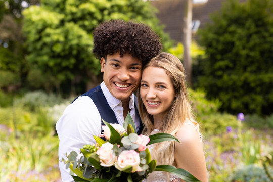 Portrait of smiling diverse bride and groom holding bouquet in garden at outdoor wedding