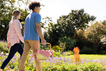 Happy diverse couple holding hands walking in sunny garden with picnic basket, with copy space