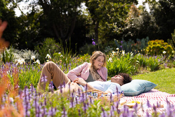 Happy diverse couple lying on blanket, having picnic, talking in sunny garden
