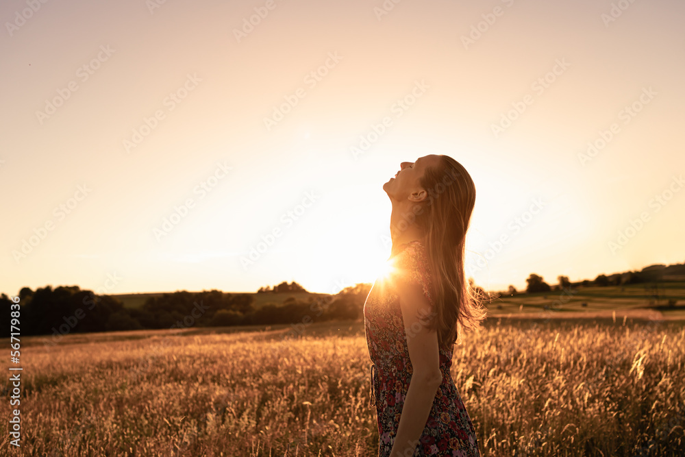 Wall mural Joyful young beautiful woman standing in a field at sunset looking up to the sky feeling happy, hopeful and free in nature  