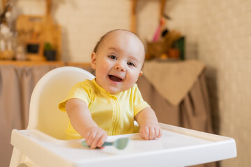 a cute baby in a yellow bodysuit is sitting in a high chair in the kitchen of the house with a plastic spoon. Baby food, first complementary food