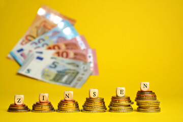 German word for interest charges, ZINSEN, from wood cubes on stacks of coins. Euro cash money on yellow background, coins and banknotes, close up