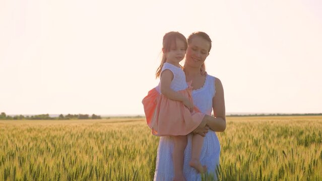 Mom little daughter are walking in green wheat field, hugging and kissing. Child mom are walking in wheat field. Happy family travel. Baby is in moms arms. Farmer woman child walk in field. Kid mom