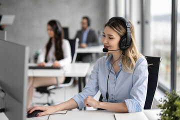 Young blonde woman in headset, sitting in office, working with her colleagues in customer support service.