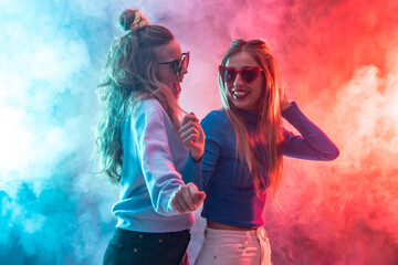 Two young blonde caucasian women dancing in nightclub, dancing having fun at party, red and blue led smoke