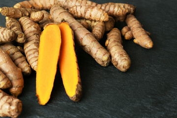 Whole and cut turmeric roots on black textured table, closeup