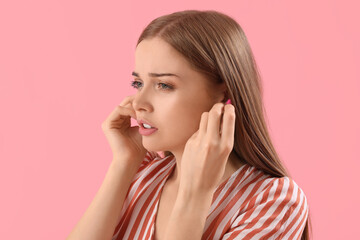 Young woman with ear plugs suffering from loud noise on pink background, closeup