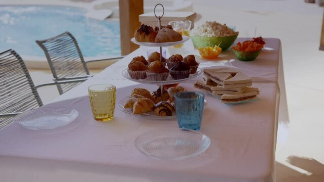 Table with sweets for a poolside party in summer, with a tray with croissants, sandwich, popcorn, crudit&eacute;s, coloured glasses, transparent plates and white tablecloth.