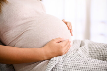 Asian pregnant woman in casual clothes relaxing on sofa in her living room, touching her belly