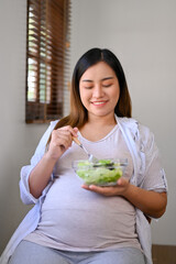 Happy Asian pregnant woman eating a healthy salad for breakfast at a table by the window.