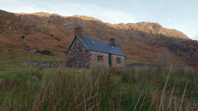 Rising slowly from the vegetation in the foreground reveals an old stone building (a Scottish Bothy called Glendhu) at the base of a mountain in a remote glen in rural Scotland.