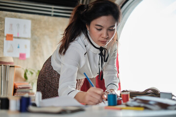 Fototapeta premium Asian Thai young teenage female fashion designer works in studio by idea drawing sketches and craft fabric for a creative dress design collection jobs, professional boutique tailor SME entrepreneur.