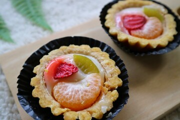 Kue pia buah or Small fruit pie cake on a black round plate and wooden pad with orange amd strawberry on the top. Used to called Kue Pia Buah. White fluffy background.