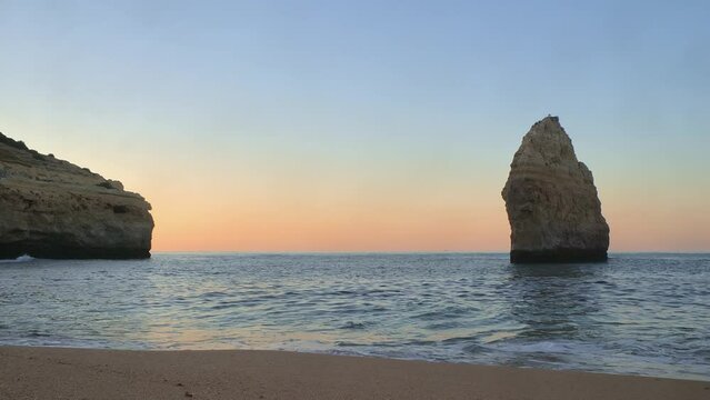 Ground level footage of gentle waves lapping on a beautiful deserted beach (Praia do Carvalho) in Algarve, Portugal at sunrise showing sandstone cliffs and a sea stack.