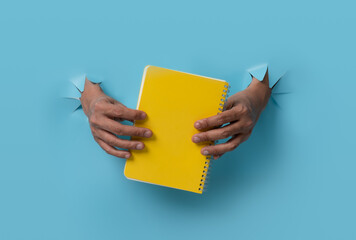 Male hand holding a book through blue paper background.
