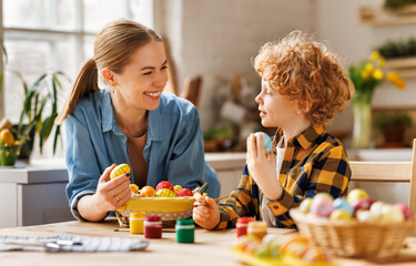 Loving young mother teaching happy little kid son to decorate Easter eggs while sitting in kitchen