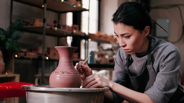 Woman potter uses ribbon tool with which makes pattern on clay vase, closeup. Craftsman in workshop to apply draws a picture on clay vase. Creativity and traditional crafts concept