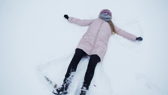 Teenage girl in pink jacket making snow angels in a cold winter day. 